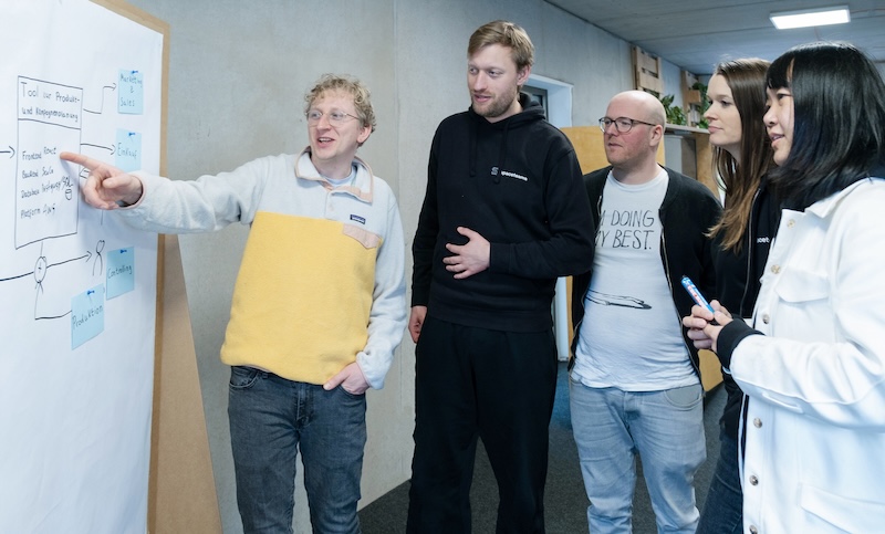 Five people are standing next to a pin board with a big sheet of paper and some blue sticky notes. One person is pointing at some text that is written on the papers and explains something to the others.