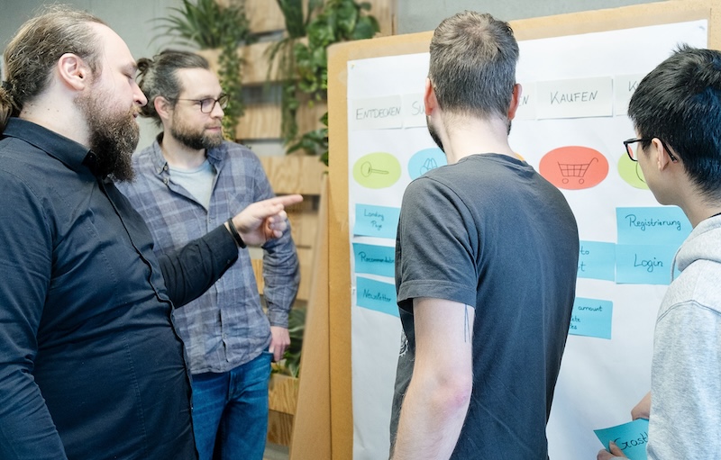 Four people standing in front of a pin board. One person is pointing at the board. On the board we see colorful sticky notes with words and symbols about a webshop.