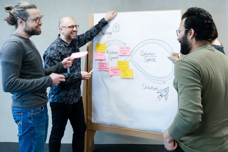 Four people standing next to a pin board, one is holding a pen, one is holding a colored card. On the board we see a drawing about 